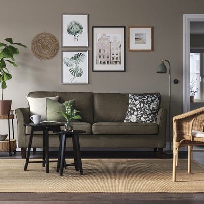 Hyggelig stue med EKHOLMA sofa, grønne puder, galleri vægkunst, kurvestol og sidebord med krus og plante.