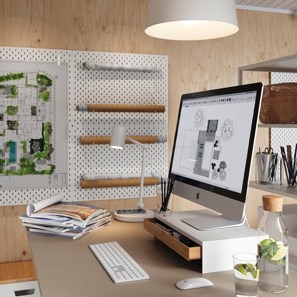 Hjemmekontor skrivebord med imac, tastatur, mus, lampe og bøger. Pegboard med tegninger og grafik. Hvid og træ indretning.