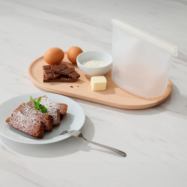 Træbakke med æg, chokolade, sukker, smør og en klar silikone FILFISK-pose . Nedenunder holder en hvid tallerken kagestykker med en sølvgaffel.