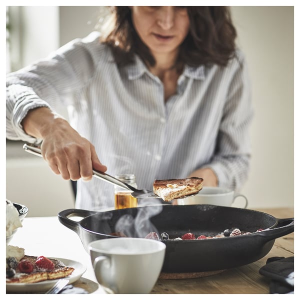 En person, der bruger en FINMAT-spatel i rustfrit stål, der holder en tilberedt madskive over en stegepande med bær, på et træbord med en kop kaffe.