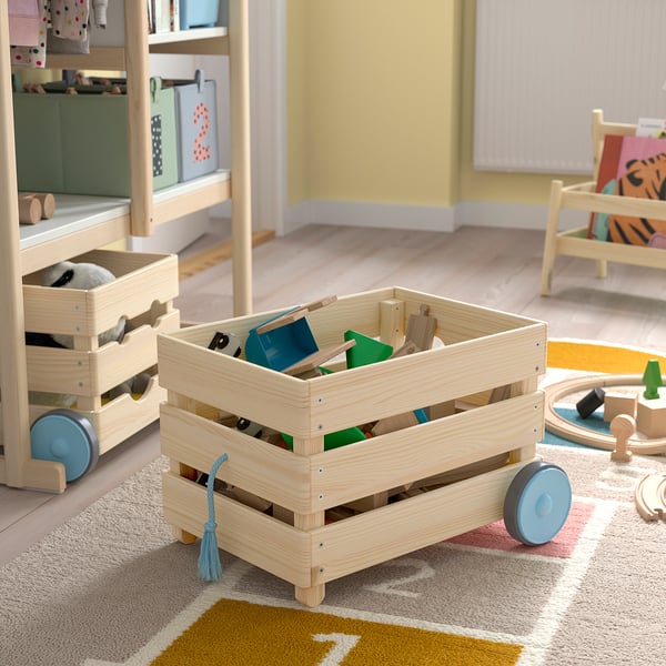 En trælegetøjsopbevaringsvogn på hjul fyldt med børnelegetøj, placeret på et farverigt tæppe i et lyst, legende rum.