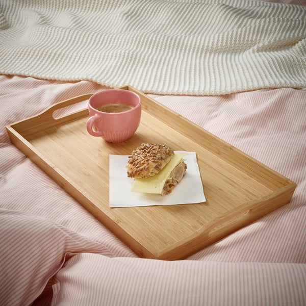 Morgenmadsbakke i træ på en seng med lyserøde lagner, der holder en kop kaffe og en sandwich, hvilket giver en hyggelig morgenmadsoplevelse.