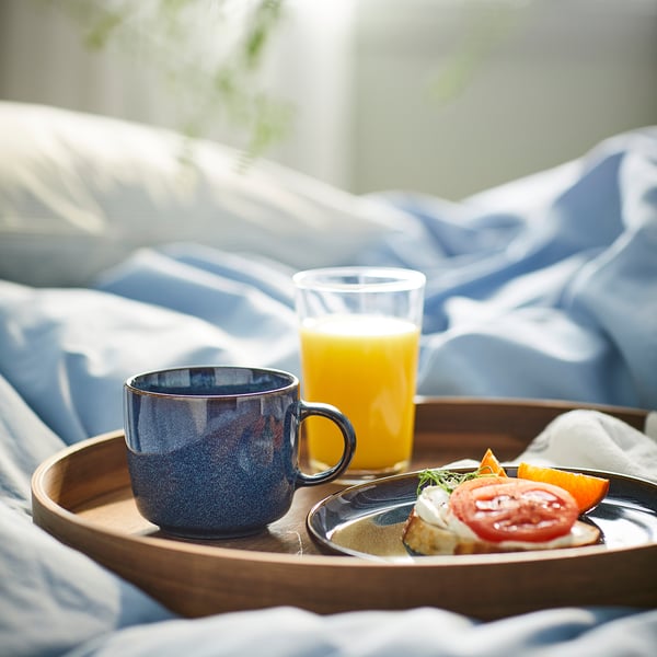 Blåt stentøjskrus, appelsinjuice, toast på træbakke mod blåt sengetæppe.