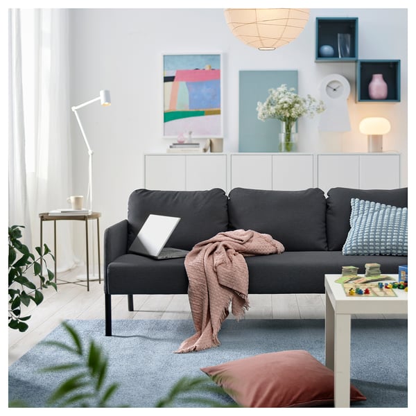 Moderne stue med sort sofa, gråt tæppe, blå puder, bærbar computer, tæppe, sofabord, lampe, plante og farverig kunst.