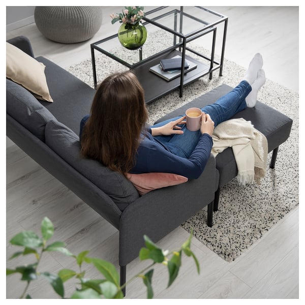 Kvinde på GLOSTAD sofa med kaffe, hyggeligt tæppe og blomster.