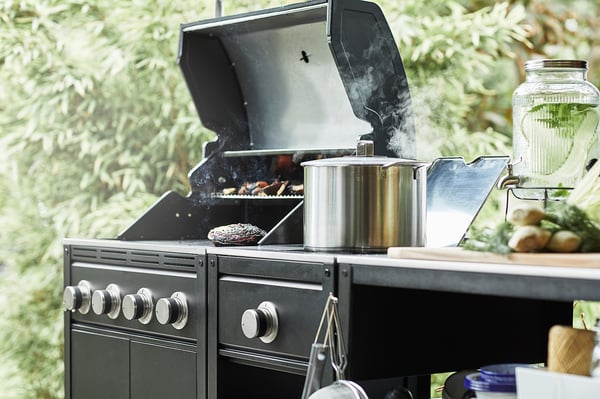 Udekøkken med gasgrill og sidebrænder; røg stiger, gryder i rustfrit stål, grøn baggrund.
