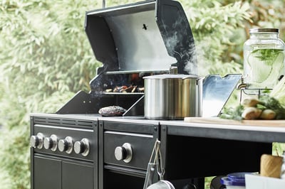 Udekøkken med gasgrill og sidebrænder; røg stiger, gryder i rustfrit stål, grøn baggrund.