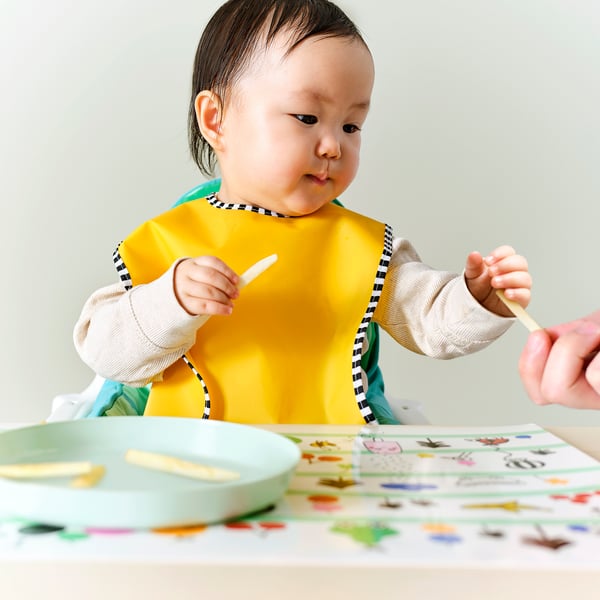 Baby iført gul hagesmæk, holder ske, voksen hånd tilbyder en anden. Bordet har farverige sticky notes.