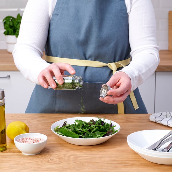 En person i et forklæde krydrer en salat med urter fra en lille glaskrukke, omgivet af citroner, salt, olie og redskaber på en bordplade af træ.