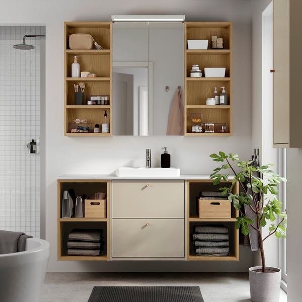 Moderne badeværelsesvask i beige, HAVBÄCK, med træhylder, keramisk vask og spejl. Planter, håndklæder og toiletartikler er synlige. Vægmonteret, rummeligt.