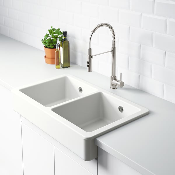 En moderne køkkenvask med en hvid dobbeltvask og et slankt armatur med en grøn plante og to glasflasker ved siden af.