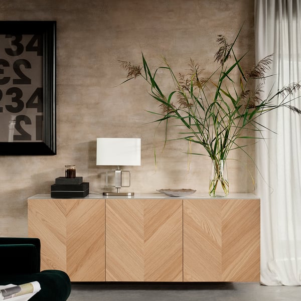 Minimalistisk opholdsrum med moderne lyst egetræsskab, hvid lampe, sorte genstande, høj plante i vase, grøn fløjlsstol, abstrakt kunst på væggen.