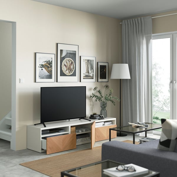 Moderne stue med tv på egetræsstativ, indrammede billeder og sofabord i glas foran grå sofa.