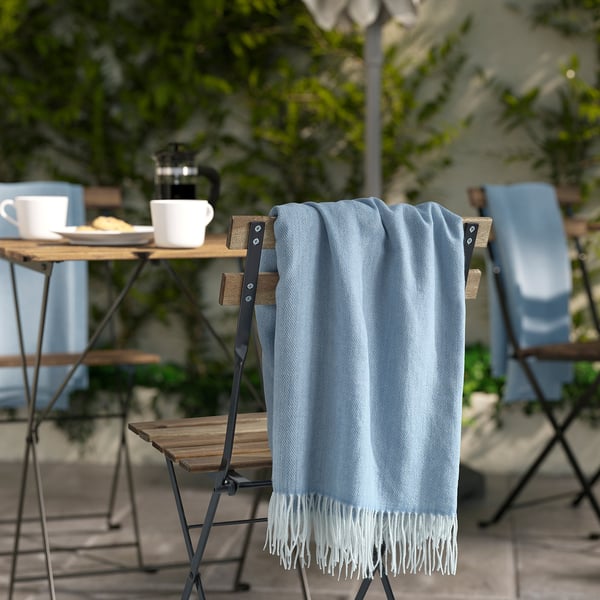 Lyseblå HOLMVI-plaider med hvide frynser draperet på en stol, nær et træbord med sofaopstilling, på en udendørs terrasse.