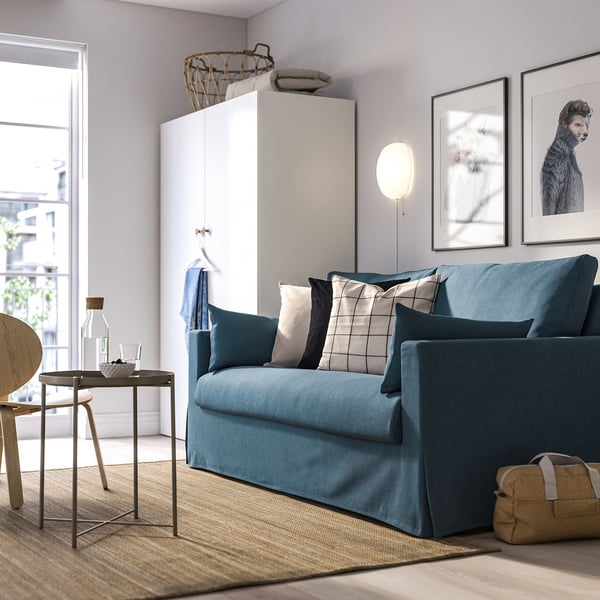 Moderne blå sofa med farverige puder i lys stue. Stor, komfortabel, med vindue og kunstværker bagved.