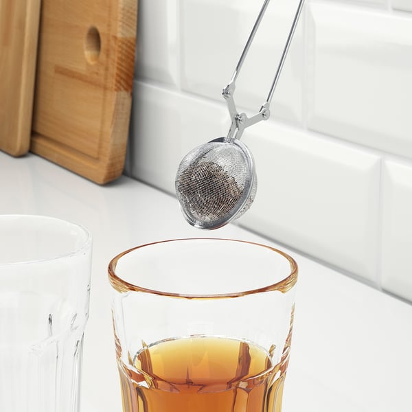 Sølv te infusionsenhed holdt af en tang dyppes i et klart glas gylden te ved siden af matchende kande.