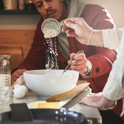 Person hælder mel i røreskålen ved hjælp af målebæger. Skål, ingredienser og køkkenredskaber er på bordet.