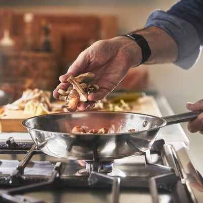 En person laver mad ved at tilføje svampe til en sydende pande på en gaskomfur.