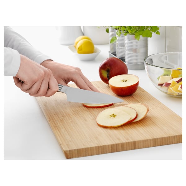 Hænder skærer æble på ikea knivbræt af træ, rustfri kniv, skål med frugt i nærheden.