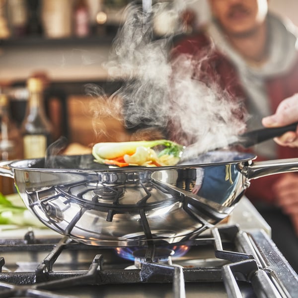 I en wok i rustfrit stål med non-stick belægning tilberedes farverig mad på et gaskomfur, der udsender damp.