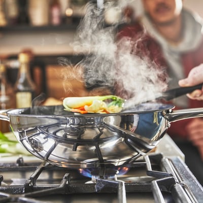I en wok i rustfrit stål med non-stick belægning tilberedes farverig mad på et gaskomfur, der udsender damp.
