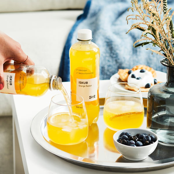 Hånden hælder orange ISKUB-drik i et glas sammen med et andet glas, en flaske, is og en skål med blåbær på en bakke.