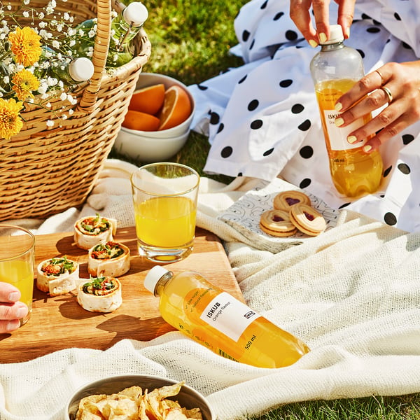 Picnicscene med appelsindrik ISKUB og snacks på et tæppe. Personen holder en flaske af drikken.