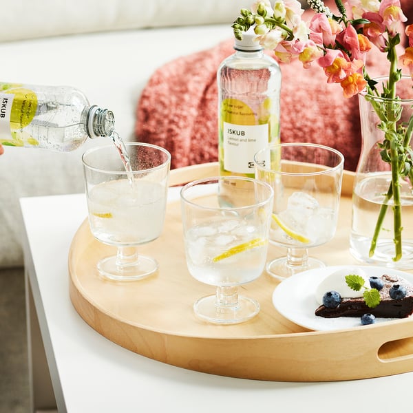 Håndhældning af ISKUB-drik med is og citron i glas. Bakken har glas, ISKUB-flaske, blåbær og blomster.
