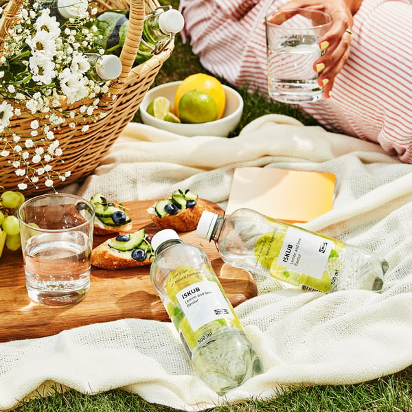 En picnicscene med en flaske ISKUB citron-lime-drik, glas vand, frisk frugt og snacks, lagt ud på et tæppe med blomster.