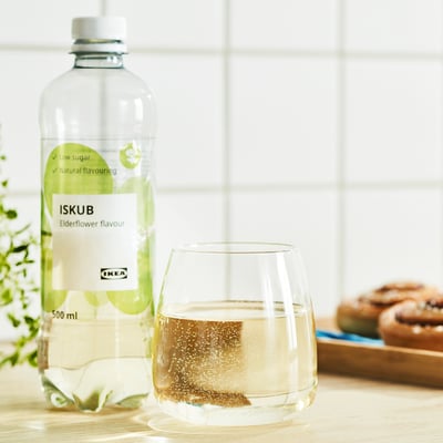 En klar flaske ikeas ISKUB hyldeblomstsmag står ved siden af et fyldt glas på et bord, omgivet af kager og grønt.