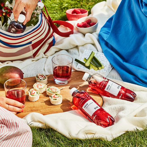 Picnicscene med ISKUB-drinks, sandwichruller, frugt og snacks lagt ud på et tæppe. Nogen hælder ISKUB-drik i et glas.