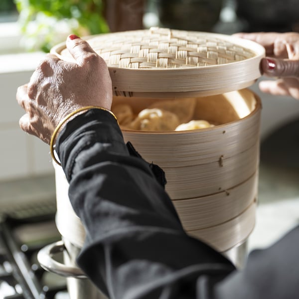 Person, der holder bambusdampkurv med dumplings, iført sort handske.