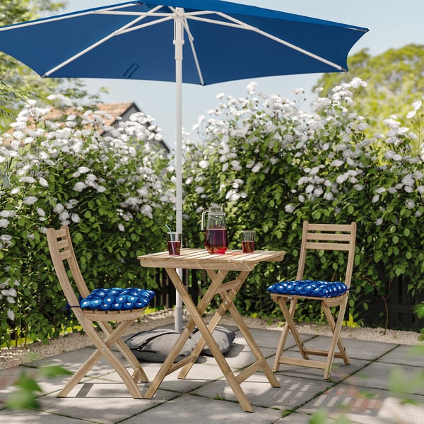 Afslappet udendørs terrasse med blå prikkede puder på træstole, blå paraply, hvide blomster, drikkevarer på træbord.