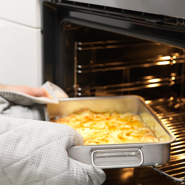 En person, der bærer ovnhandsker, fjerner en bradepande fyldt med mad fra en ovn. Dåsen har håndtag og er velegnet til både ovn og grill.