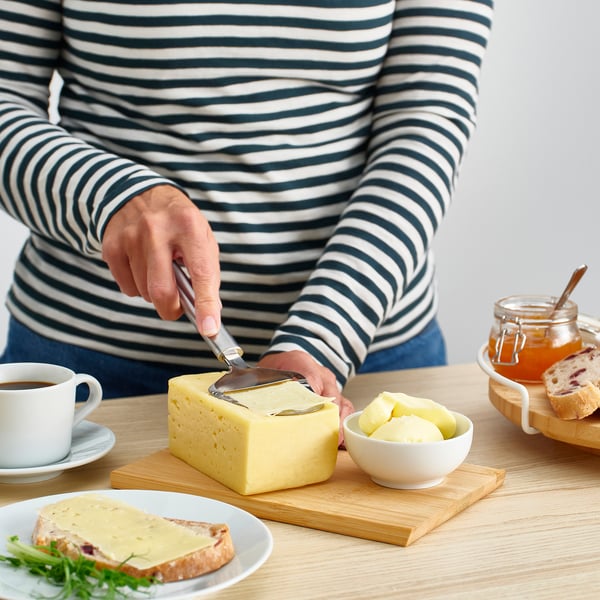 En person skærer ost på en træplade ved hjælp af en pårører i rustfrit stål. Kaffe, ost på brød og æbler er også til stede.