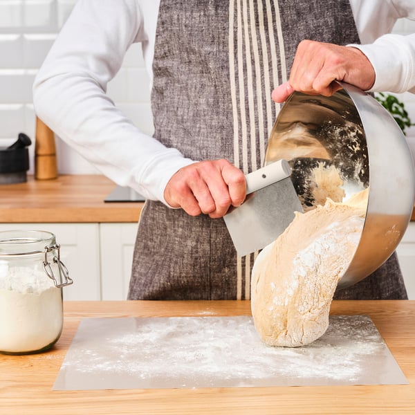 Person, der bruger en dejskærer, deler et stort stykke dej i mindre, lige store portioner på en meldrysset overflade.