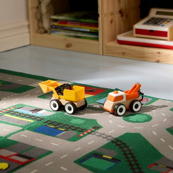 To farverige legetøjskøretøjer, gule og orange, sidder på et legetæppe med bytema og fremmer fantasifuld leg.
