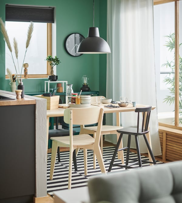 Moderne spiseplads med grønne vægge, sort loftslampe, træbord, fire lyse træstole, stribet tæppe og stort vindue med hvide gardiner.