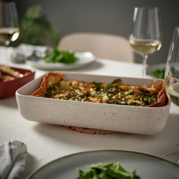 Beige LYCKAD ovnfad med mad, vinglas og tallerkener på et bord.