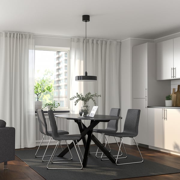 Moderne spiseplads med sort cirkulært bord og grå stole. Stort vindue med gardiner, hvide skabe. Rent, elegant design.