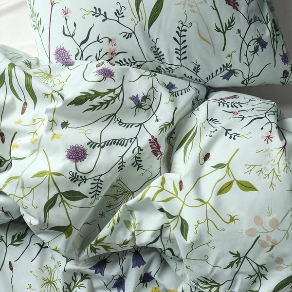Blødt, lyseblåt dynebetræk med detaljerede blomster- og grønne plantemønstre, der fremhæver bomuldstekstur af høj kvalitet og et indviklet design.