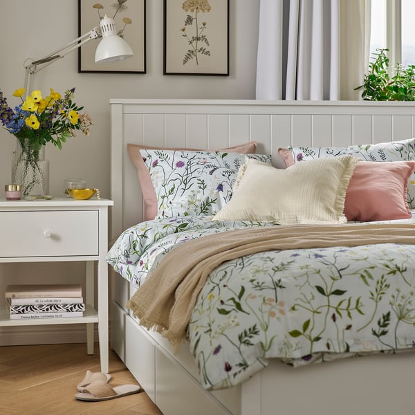 Soveværelse med MATTOXBÄR dynesæt, lyseblåt blomsterprint, hvidt hovedgavl, lyserøde puder, natbord med bøger, blomster, lampe.