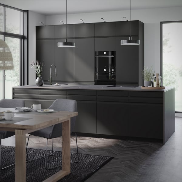 Moderne køkken med sorte skabe, hvidevarer i rustfrit stål og minimalistiske pendellamper. Spisebord i træ med stole og gråt tæppe.