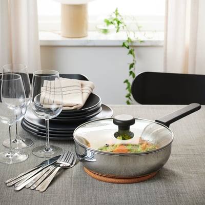 En moderne køkkenbordsopstilling med sauterpande, tallerkener, glas og bestik, klar til at servere et måltid. Gryden har et glaslåg på en bordskåner.