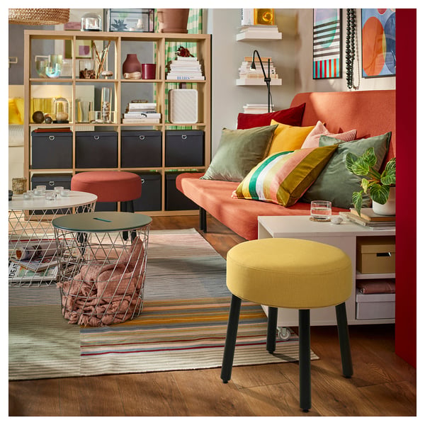 Levende stue med orange sofa, farverige puder, gul skammel, trådbord, stribet tæppe og dekorerede hylder.