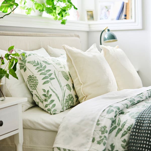 Lyst soveværelse med stor NÅLBJÖRNBÄR grøn blomsterdyne på en rummelig seng, suppleret af hvide puder, grønne planter og en markant turkis lampe.