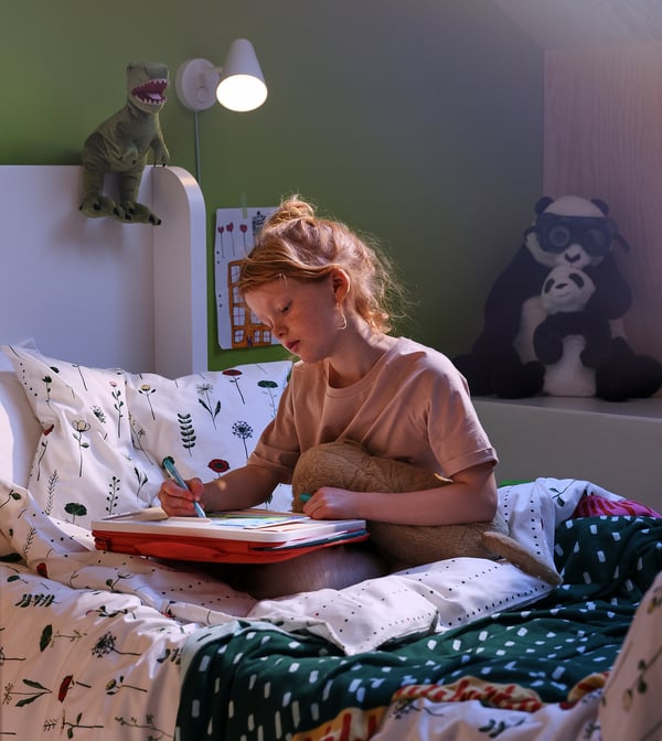 Ung pige sidder i sengen og skriver i bog. Lagen har blomsterdesign. Dinosaur lampe på hovedgærde. Panda tøjdyr i nærheden.