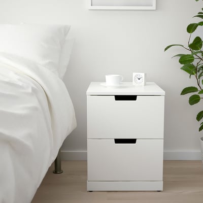 Moderne hvidt NORDLI natbord med to skuffer, mellem bed og plante.