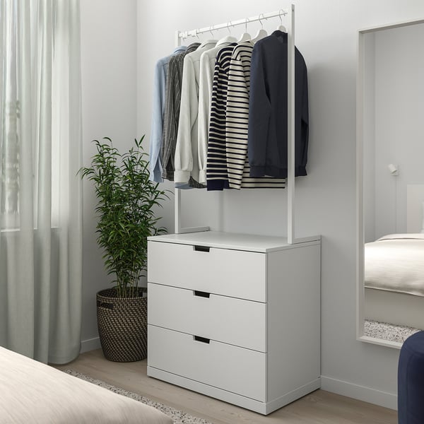 Minimalistisk soveværelse med hvid NORDLI 3-skuffers kommode under tøjstang, grøn plante ved siden af, mellem seng og vindue.