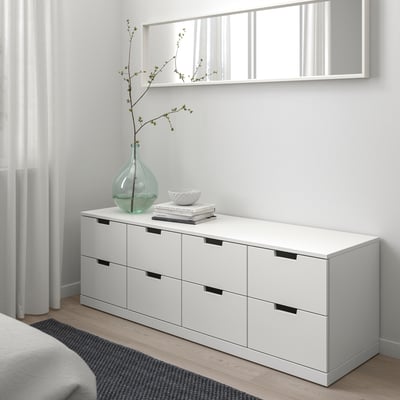 En hvid NORDLI kommode med seks skuffer, bøger, skål og vase. Rent, minimalistisk design mod en lys væg.